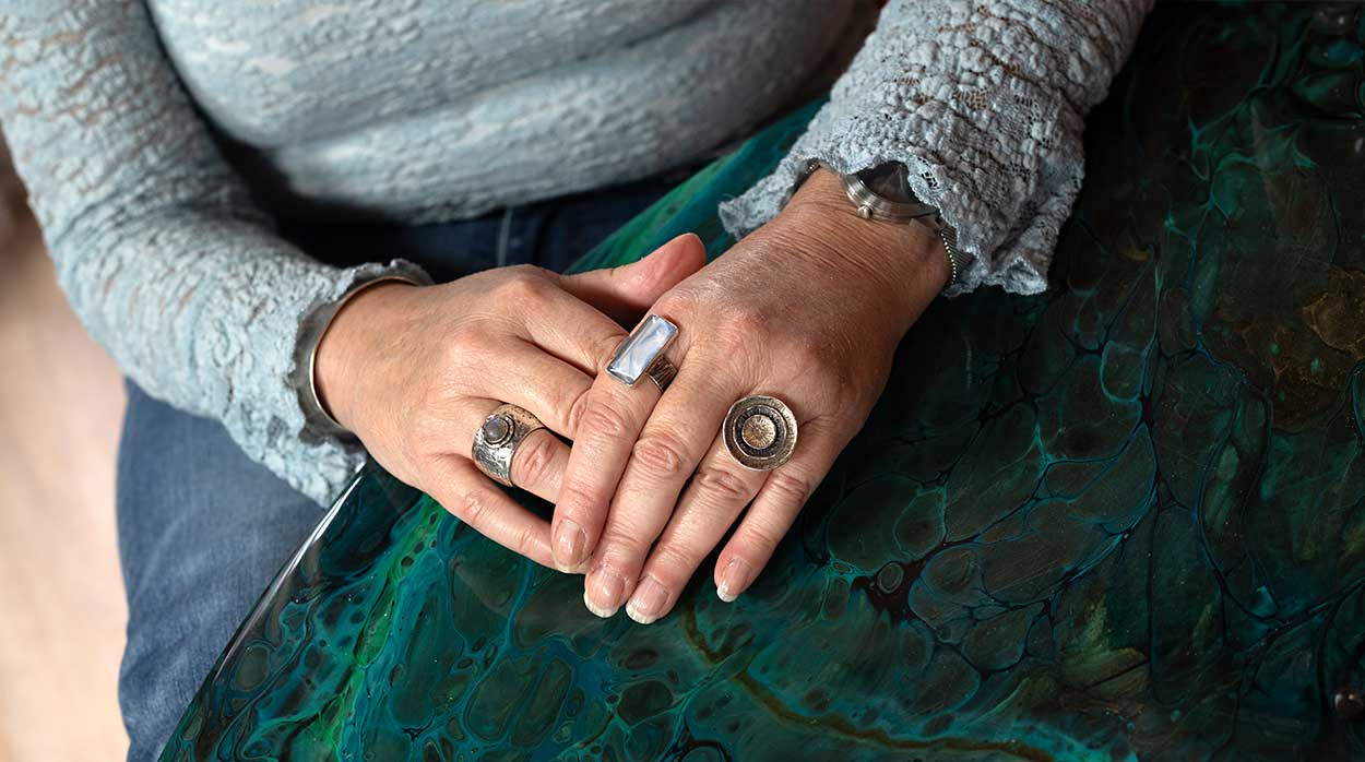 Vrouw met meerdere handgemaakte ringen aan haar vingers, rustend op een groenblauw geschilderde tafel, ter illustratie van de betekenis en symboliek van ringen combinaties bij vrouwen van Art by Daan.