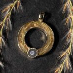Een ronde, gouden hanger met helder steentje en een stukje natuur gevangen binnenin, omringd door tere grasprieten.