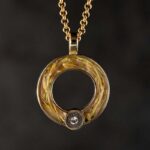 Een rond, goud hangertje met handgevlochten patroon hangt aan een ketting. Onderaan straalt een heldere steen in het warme licht.