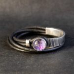 Een handgemaakte wikkelarmband van zwart leer, met een zilveren sluiting en een uniek, paars gemarmerd middenstukje.