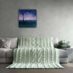 2016-Paarse-zonsopgang-50x50-interieur-e1537886644604.jpg Op de lichte grijze bank ligt een handgebreide, zachtgroene plaid en kussen. Boven de bank hangt een uniek schilderij van een paarse zonsopgang met een sierlijke boom.