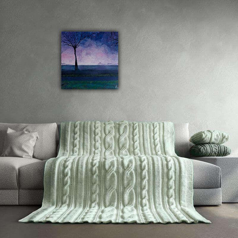 2016-Paarse-zonsopgang-50x50-interieur-e1537886644604.jpg Op de lichte grijze bank ligt een handgebreide, zachtgroene plaid en kussen. Boven de bank hangt een uniek schilderij van een paarse zonsopgang met een sierlijke boom.